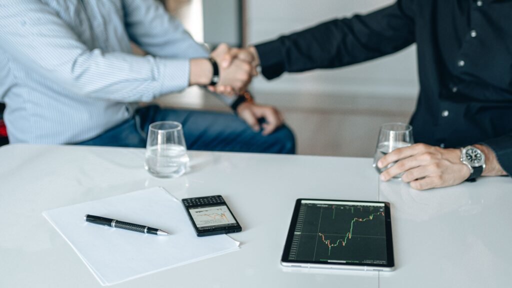 Two businesspeople shaking hands over a table with a financial chart on a tablet, representing pharma M&A and portfolio deal-making